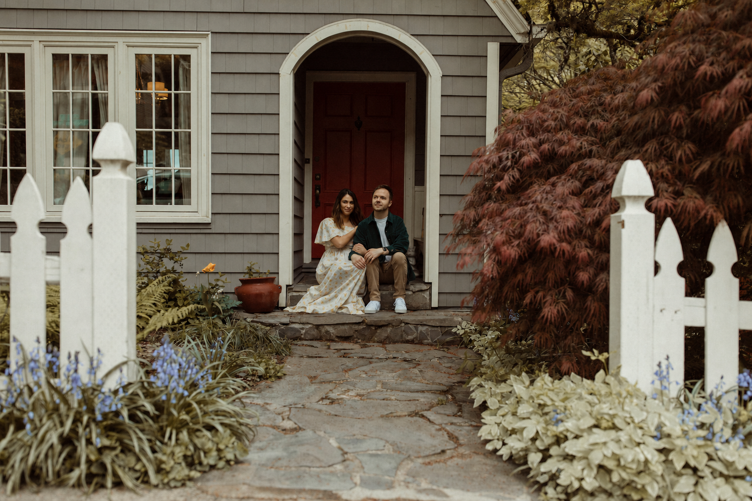 Ana and Doug sitting together on the front steps of their home, surrounded by the garden and Japanese maple